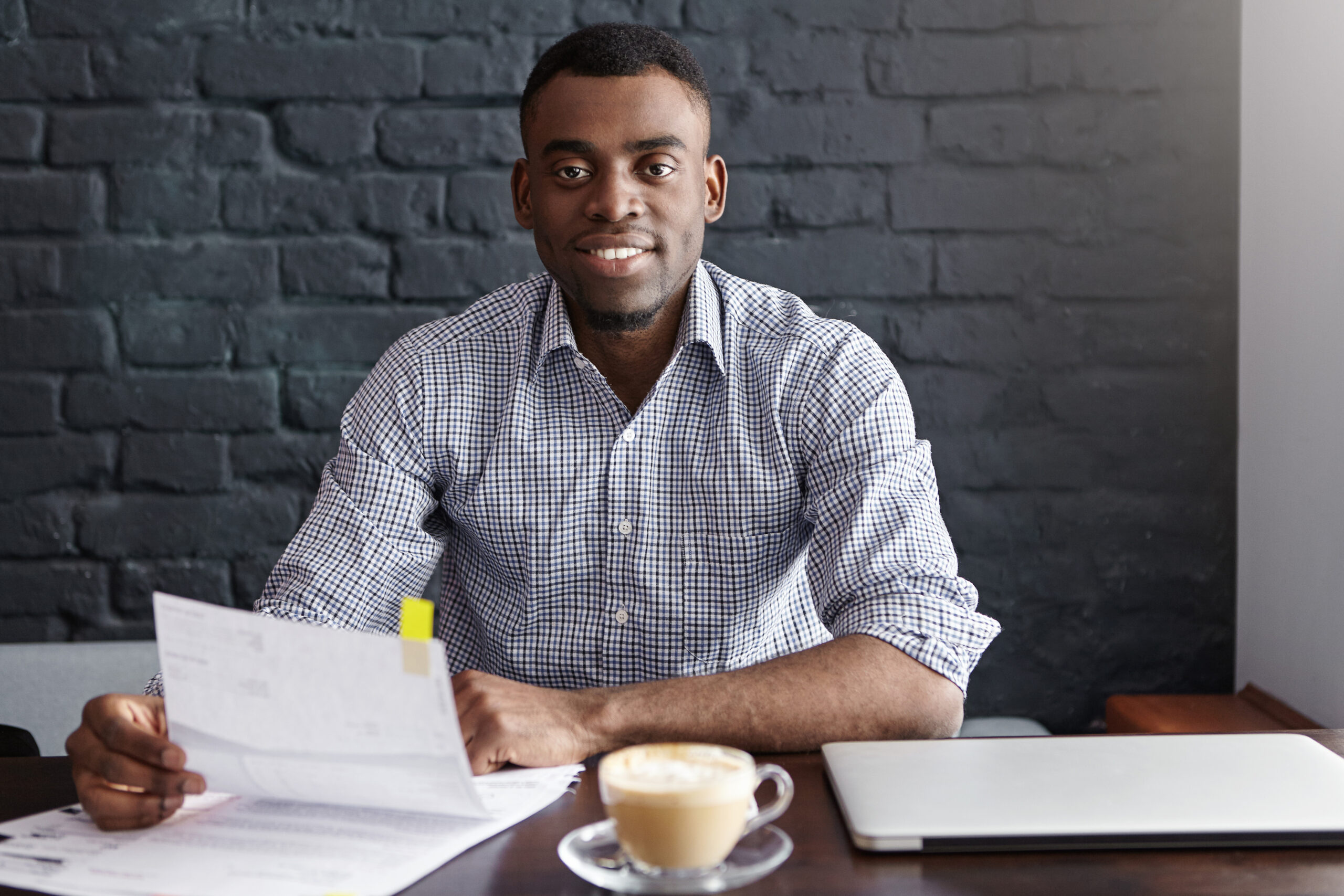 confident young african businessman wearing formal shirt sitting at table with laptop, mug and papers, holding job application during interview, recruiting people for personal assistant position